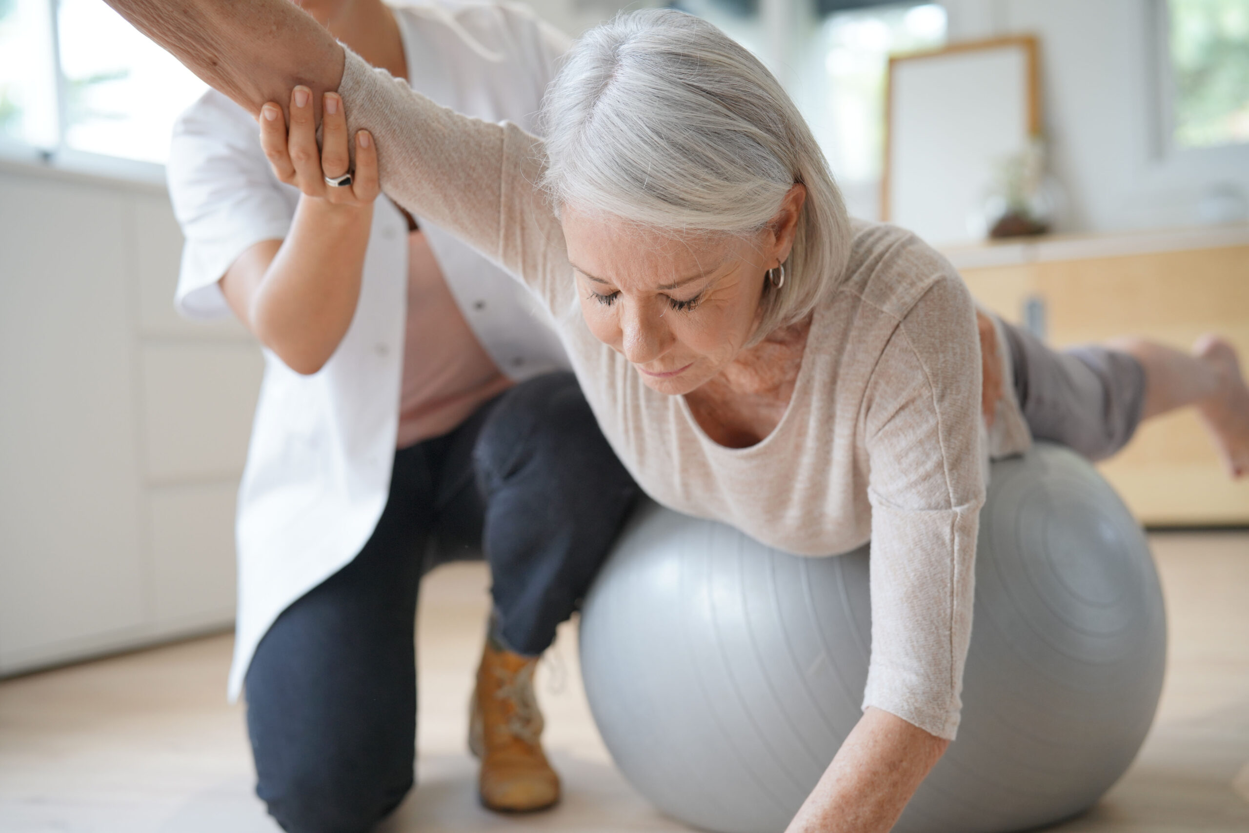 Problemas de espalda en la tercera edad: Una mujer mayor realiza con la ayuda de un fisioterapeuta ejercicios para mejorar la postura de la espalda ayudándose de una pelota de goma y apoyando su cuerpo en ella.