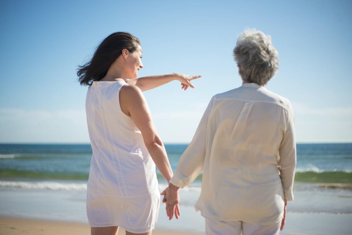 Mujer joven y mujer mayor se encuentran de espaldas a la cámara, mirando y señalando el mar