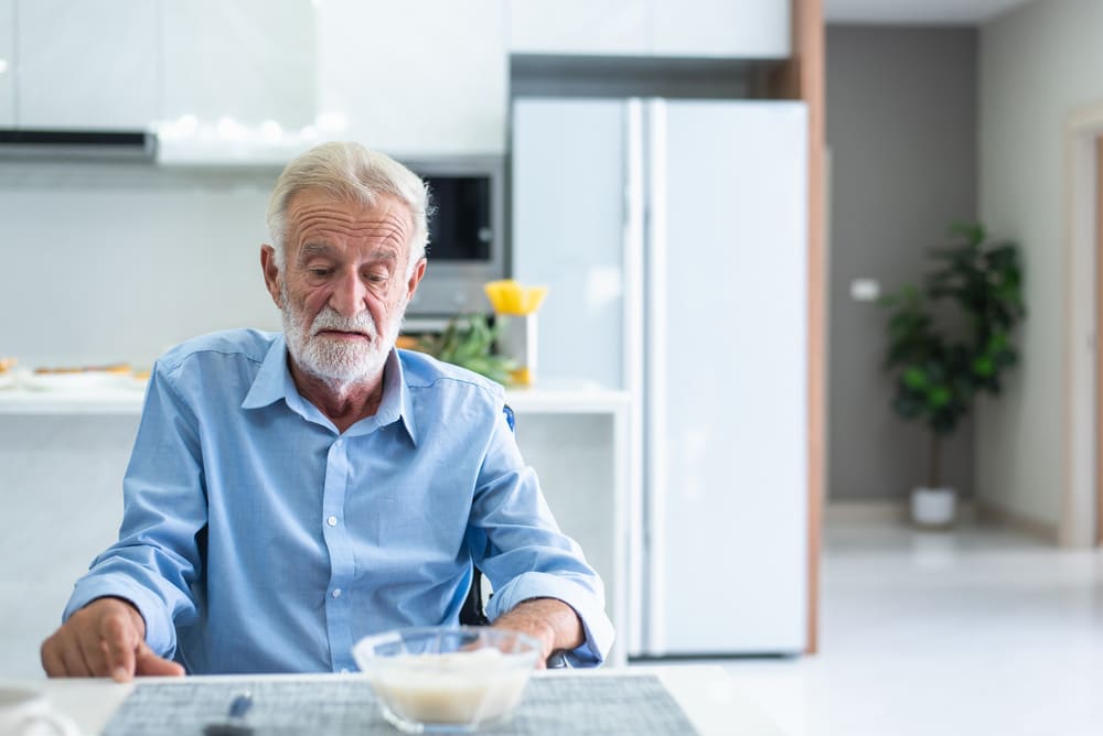 Un anciano en su casa mira un bol de arroz sentado en la mesa de la cocina. 