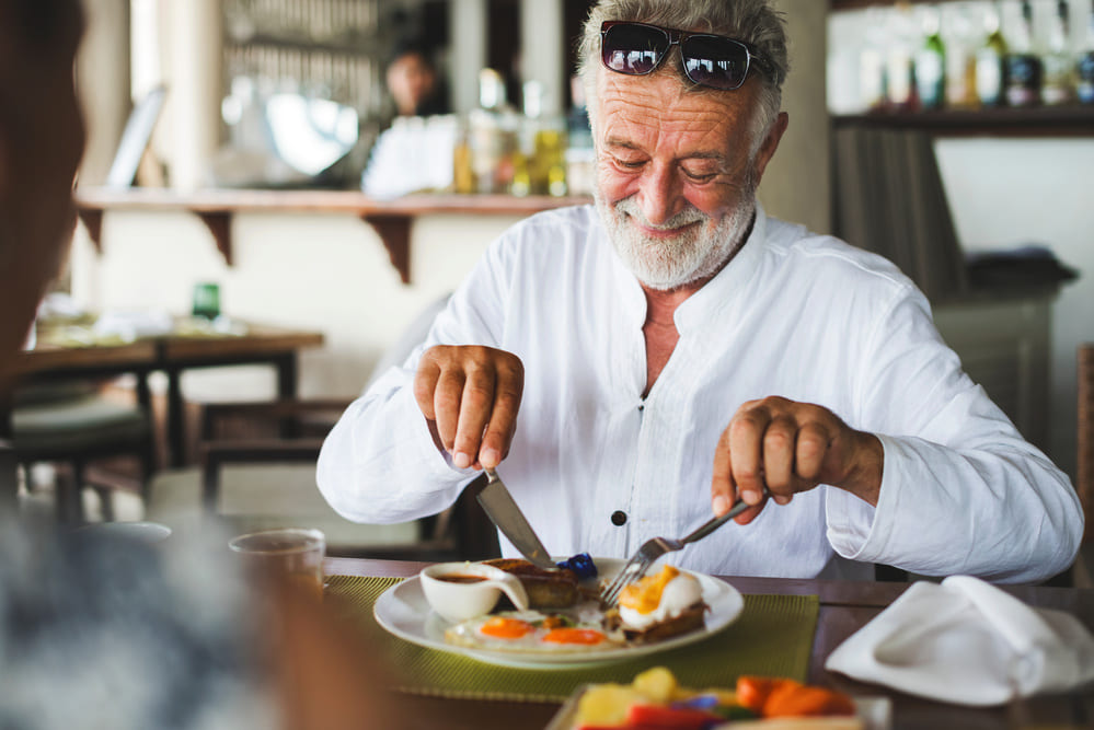 Un señor mayor corta con el cuchillo y apoyándose con el tenedor, un plato de comida que lleva huevo. 