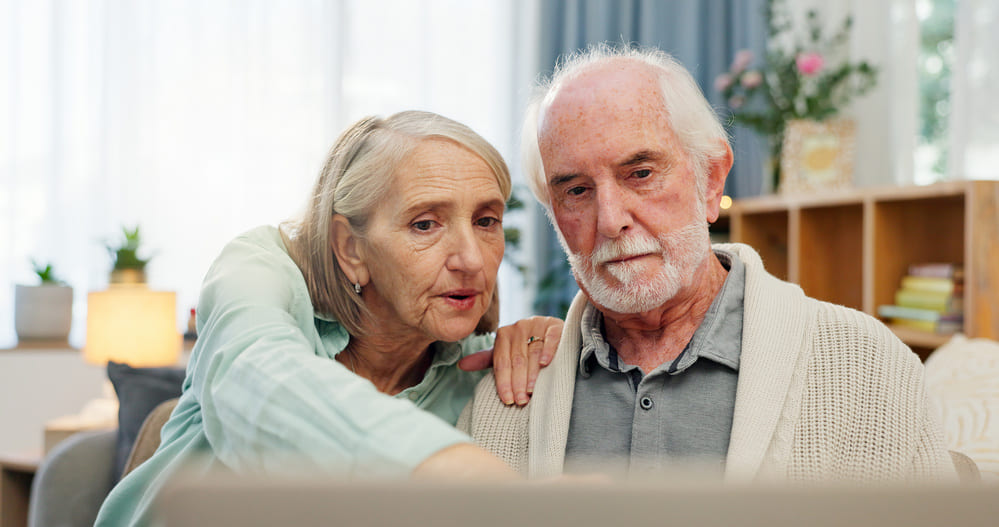 Una mujer y un hombre ancianos en el sofá de su casa miran algo en un portátil. 