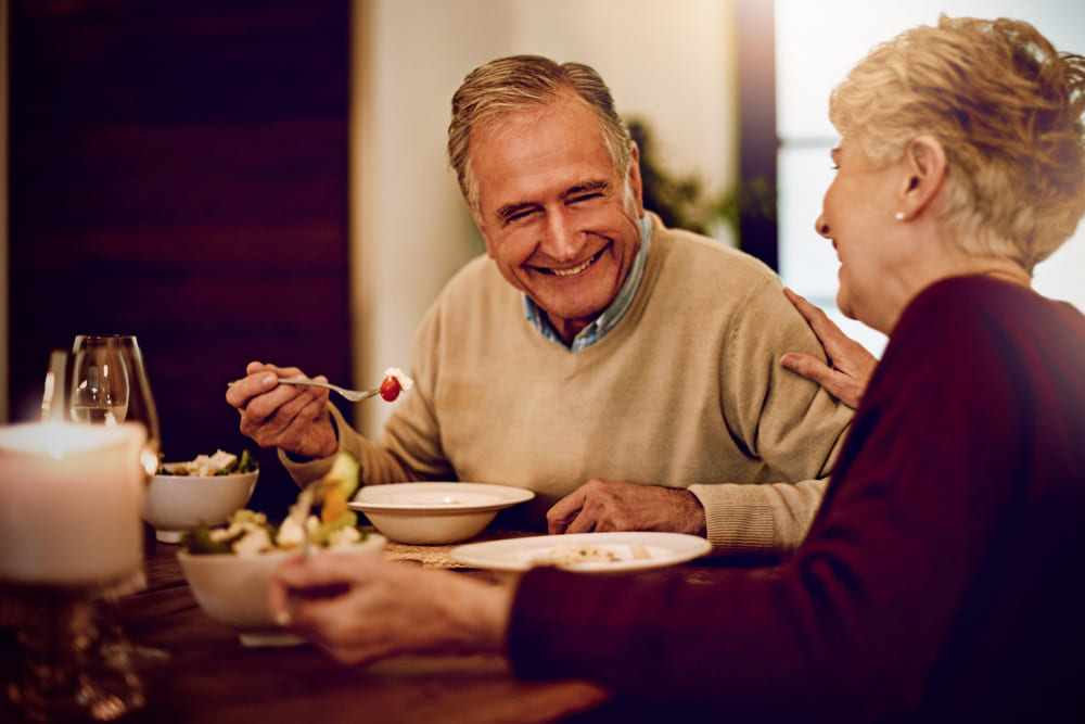 Dos ancianos cenan en una mesa con comida y el anciano porta en el tenedor un dátil. 